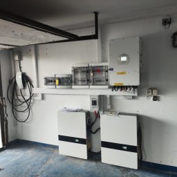 Solar-Group Sp. z o.o. - Domowa instalacja: dwa białe magazyny energii z czarnym pasem, inwerter i rozdzielnia na ścianie garażu z niebieską podłogą. Widoczny kabel do ładowania auta.