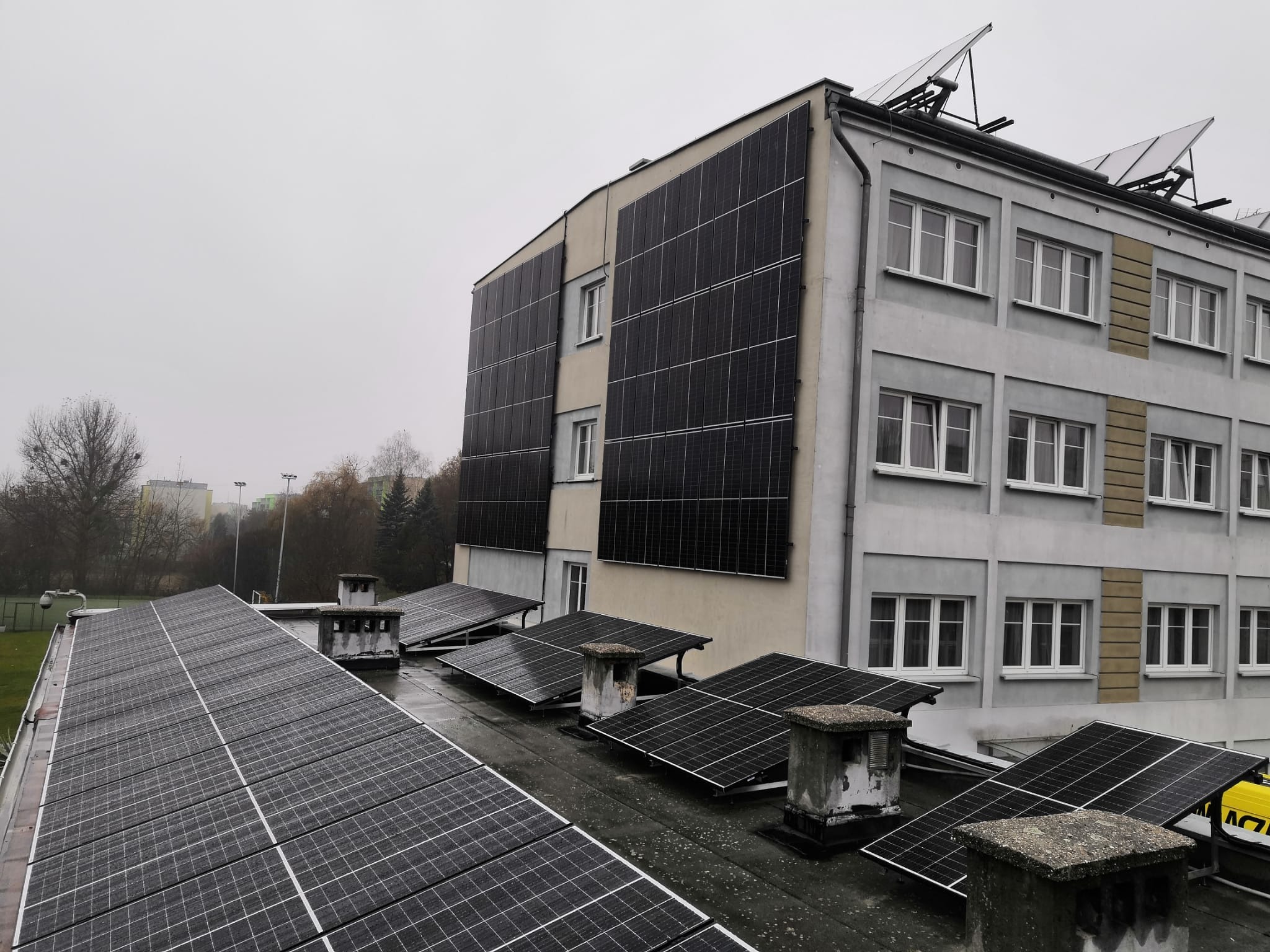 Instalacja fotowoltaiczna na dachu budynku z kominami wentylacyjnymi, widok z góry w pochmurny dzień.