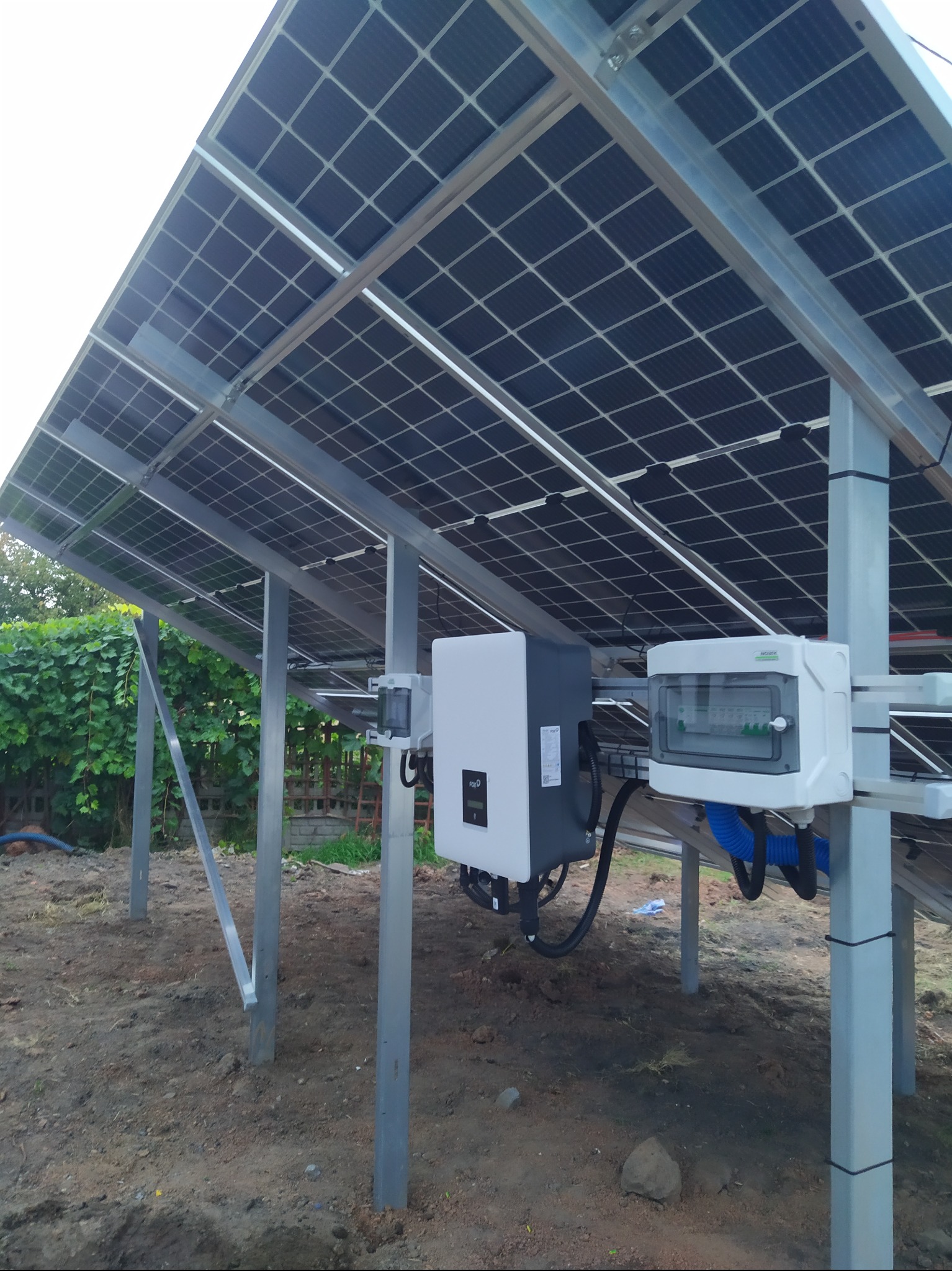 Konstrukcja wsporcza pod panele słoneczne z zamontowanym inwerterem i skrzynką zabezpieczeń na tle ogrodu.