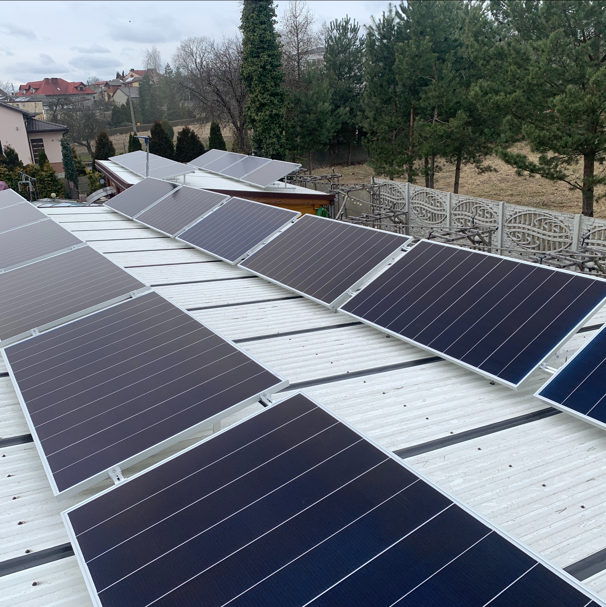 Rząd paneli słonecznych na blaszanym dachu domu jednorodzinnego w pochmurny dzień, widok z góry na instalację z widocznymi elementami mocującymi.