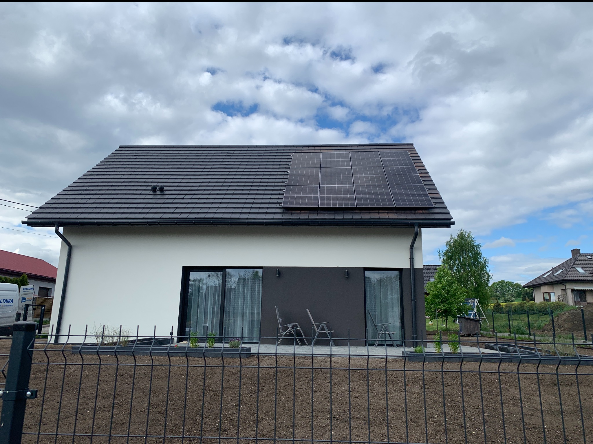 Nowoczesny dom jednorodzinny z ciemną dachówką i częściowo zamontowanymi panelami słonecznymi na dachu, widziany zza metalowego ogrodzenia.