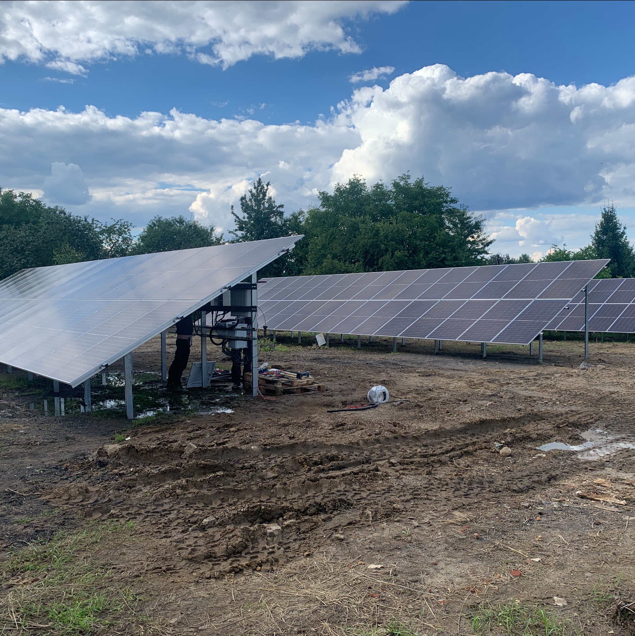 Instalacja paneli słonecznych w trakcie montażu na grząskim terenie, widoczne narzędzia i elementy montażowe, niebo z chmurami w tle.
