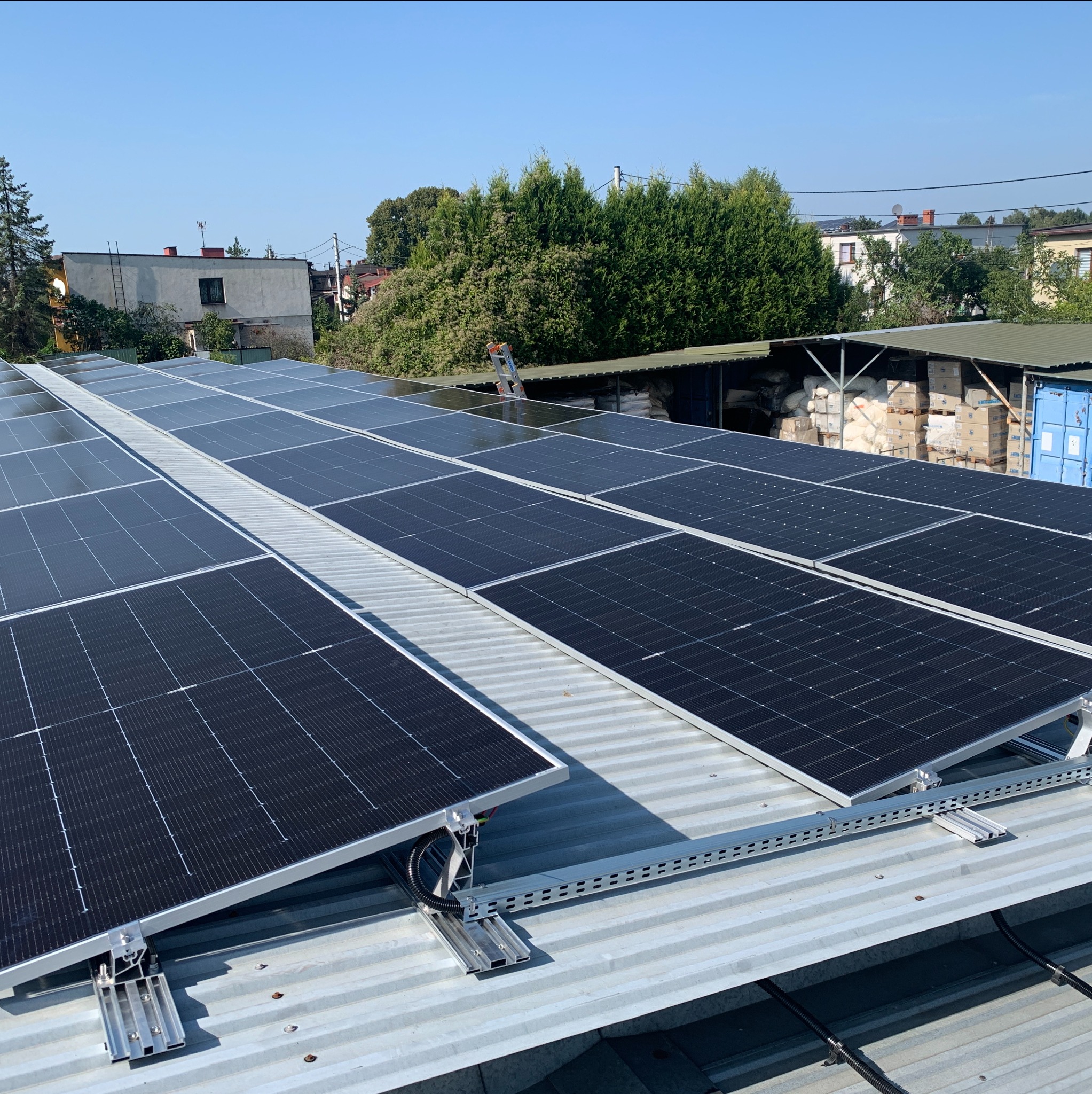 Rząd paneli fotowoltaicznych zamontowanych na metalowym dachu, widoczne metalowe konstrukcje mocujące i okablowanie, w tle budynki mieszkalne i zieleń.