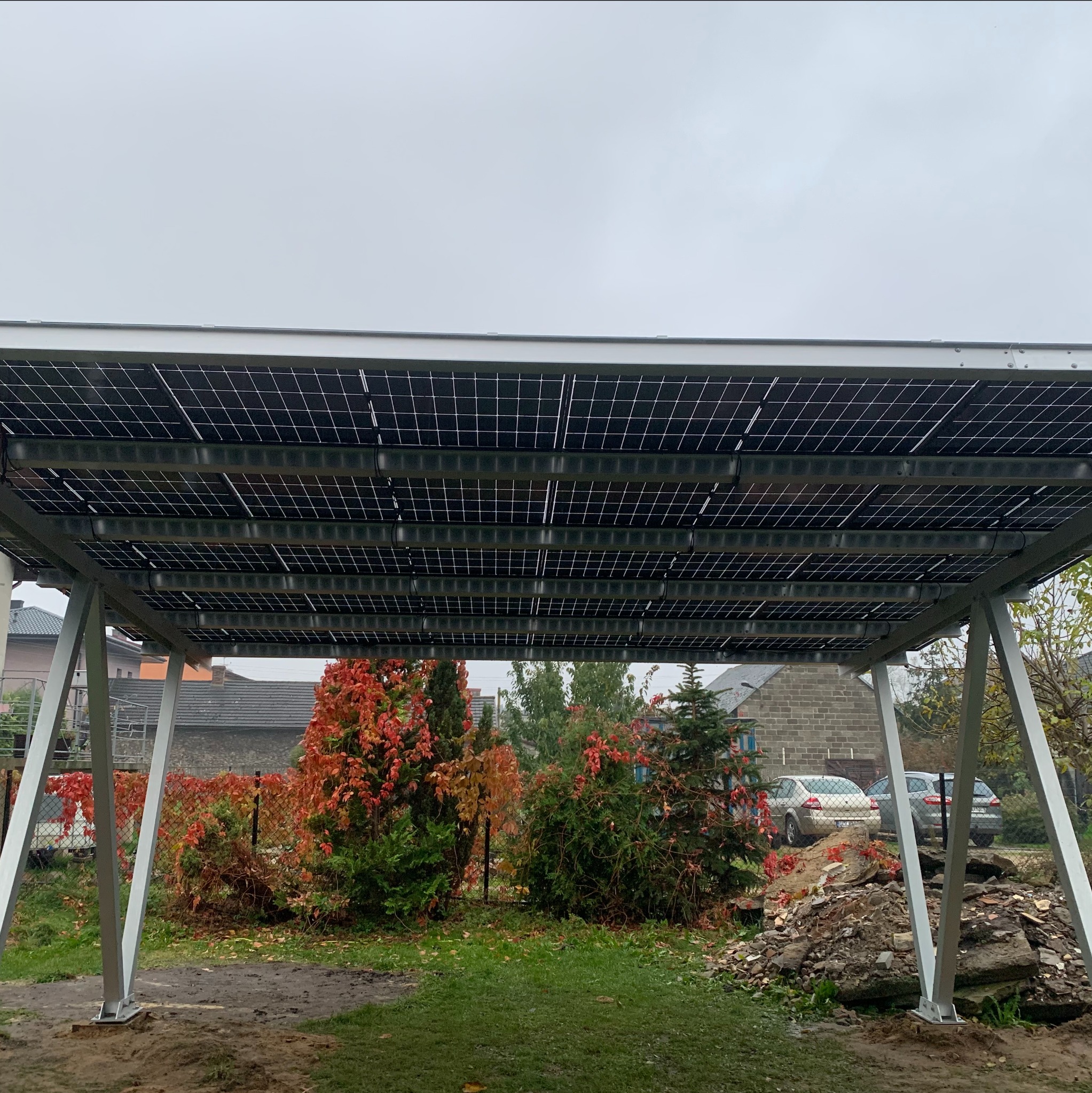 Zadaszenie carportu z panelami słonecznymi, widok od dołu, w tle zabudowa jednorodzinna i jesienna roślinność.