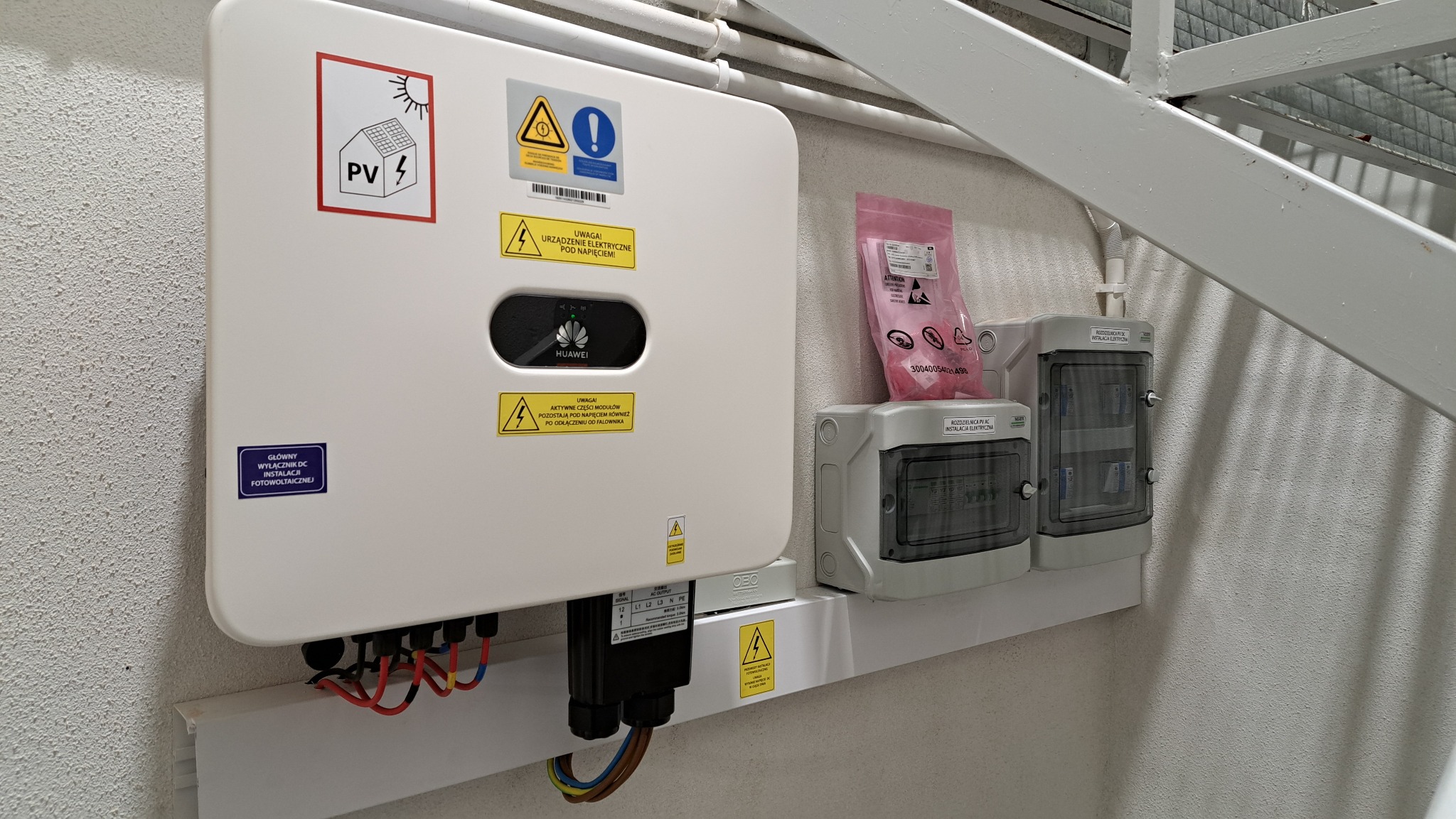 Biały falownik Huawei z naklejkami ostrzegawczymi i schematem, zamontowany na ścianie obok dwóch szarych rozdzielnic elektrycznych i pakietu akcesoriów.