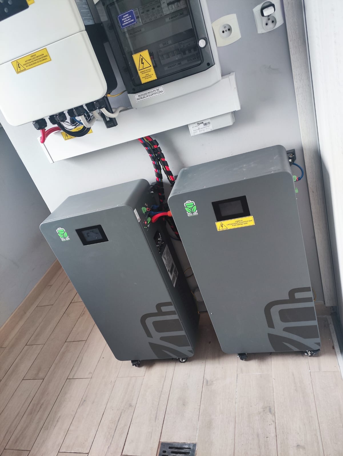 Dwa szare magazyny energii Eco BBS z podłączonymi kablami, zamontowane obok rozdzielnicy PV DC i falownika, na tle jasnej ściany i podłogi z paneli.