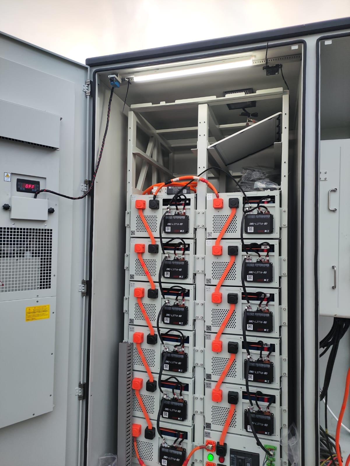 Wnętrze szafy z rzędem akumulatorów połączonych pomarańczowymi kablami. Widoczny wyświetlacz z napisem 'OFF' i elementy instalacji elektrycznej.