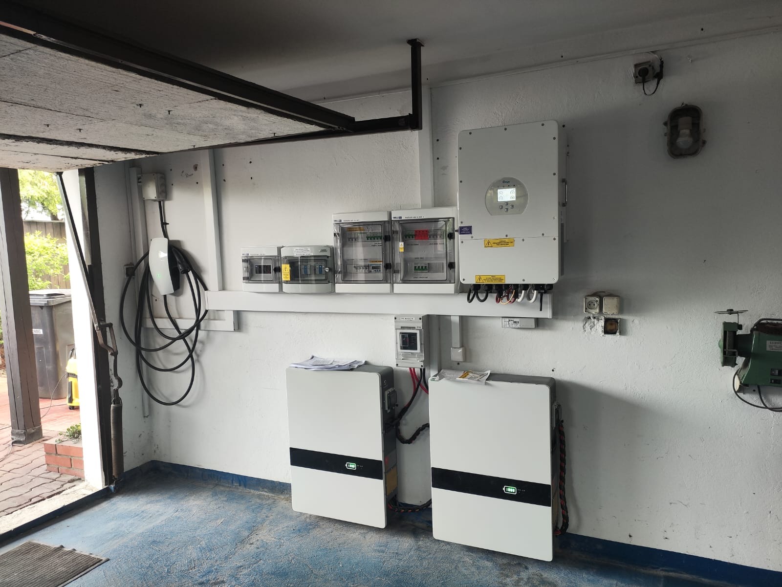 Domowa instalacja: dwa białe magazyny energii z czarnym pasem, inwerter i rozdzielnia na ścianie garażu z niebieską podłogą. Widoczny kabel do ładowania auta.