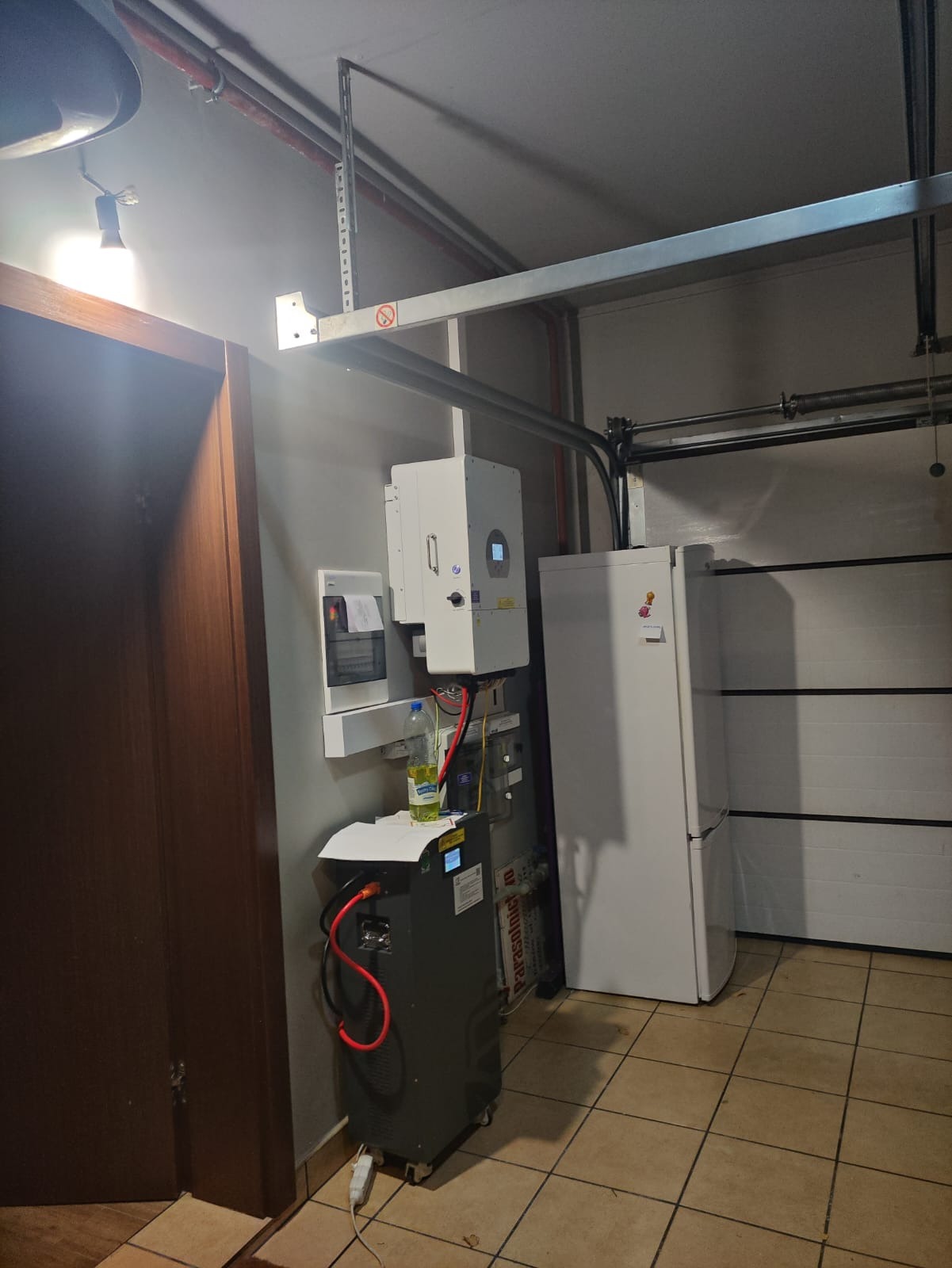 Domowa instalacja: inwerter fotowoltaiczny, magazyn energii na kółkach i skrzynka bezpiecznikowa na szarej ścianie w garażu z bramą segmentową i lodówką.