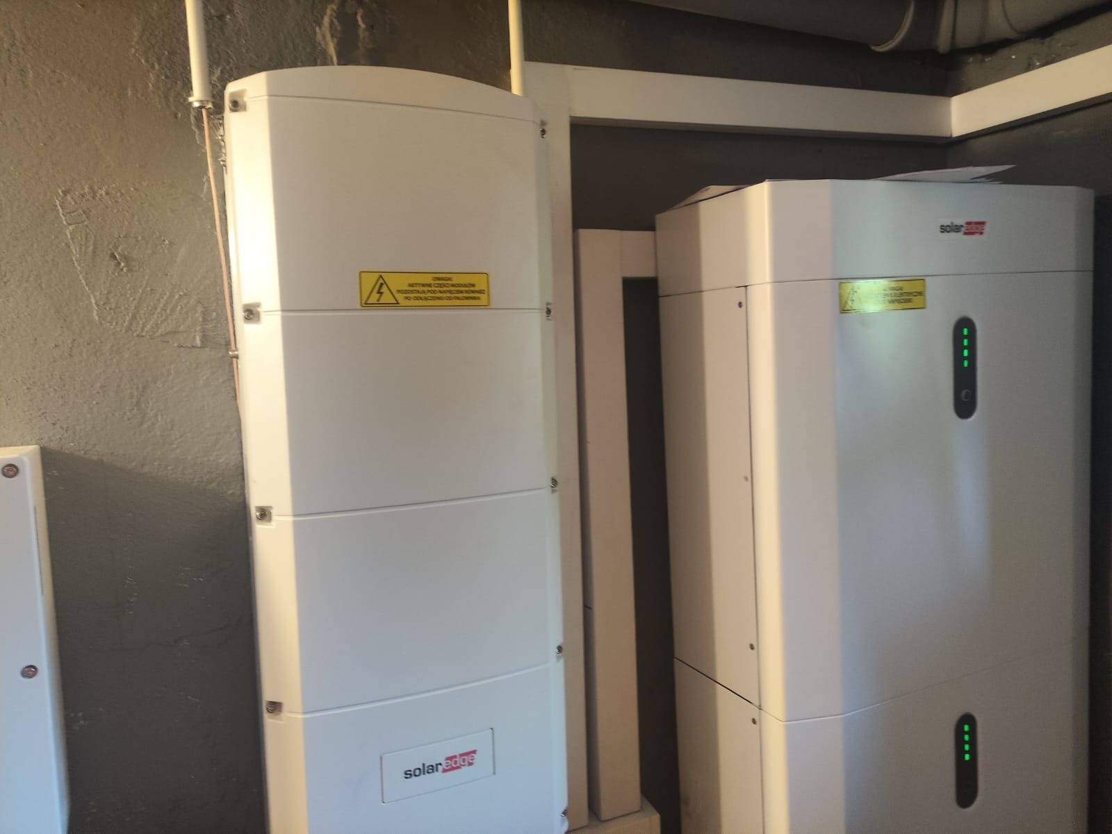 Białe moduły magazynu energii SolarEdge z widocznymi diodami LED i naklejką ostrzegawczą, zamontowane w pomieszczeniu o szarych ścianach.