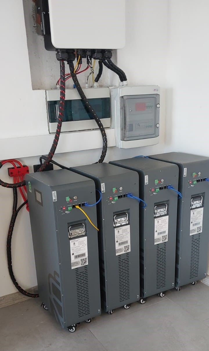 Rząd czterech szarych magazynów energii na kółkach, podłączonych kablami do białej skrzynki elektrycznej na ścianie. Widoczne kable zasilające i komunikacyjne.