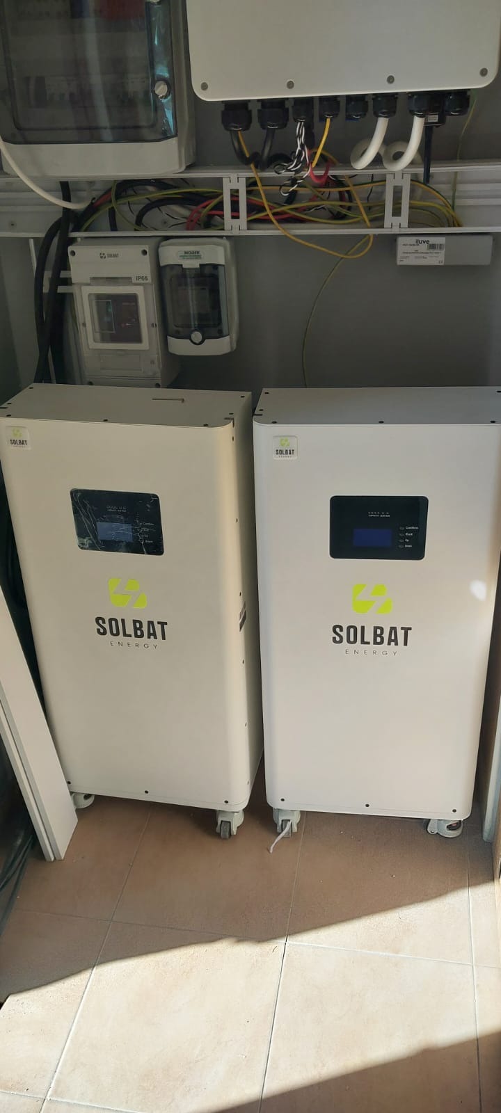 Dwa białe magazyny energii Solbat z wyświetlaczami, umieszczone na kafelkach, z widocznymi przewodami i elementami instalacji elektrycznej na ścianie.