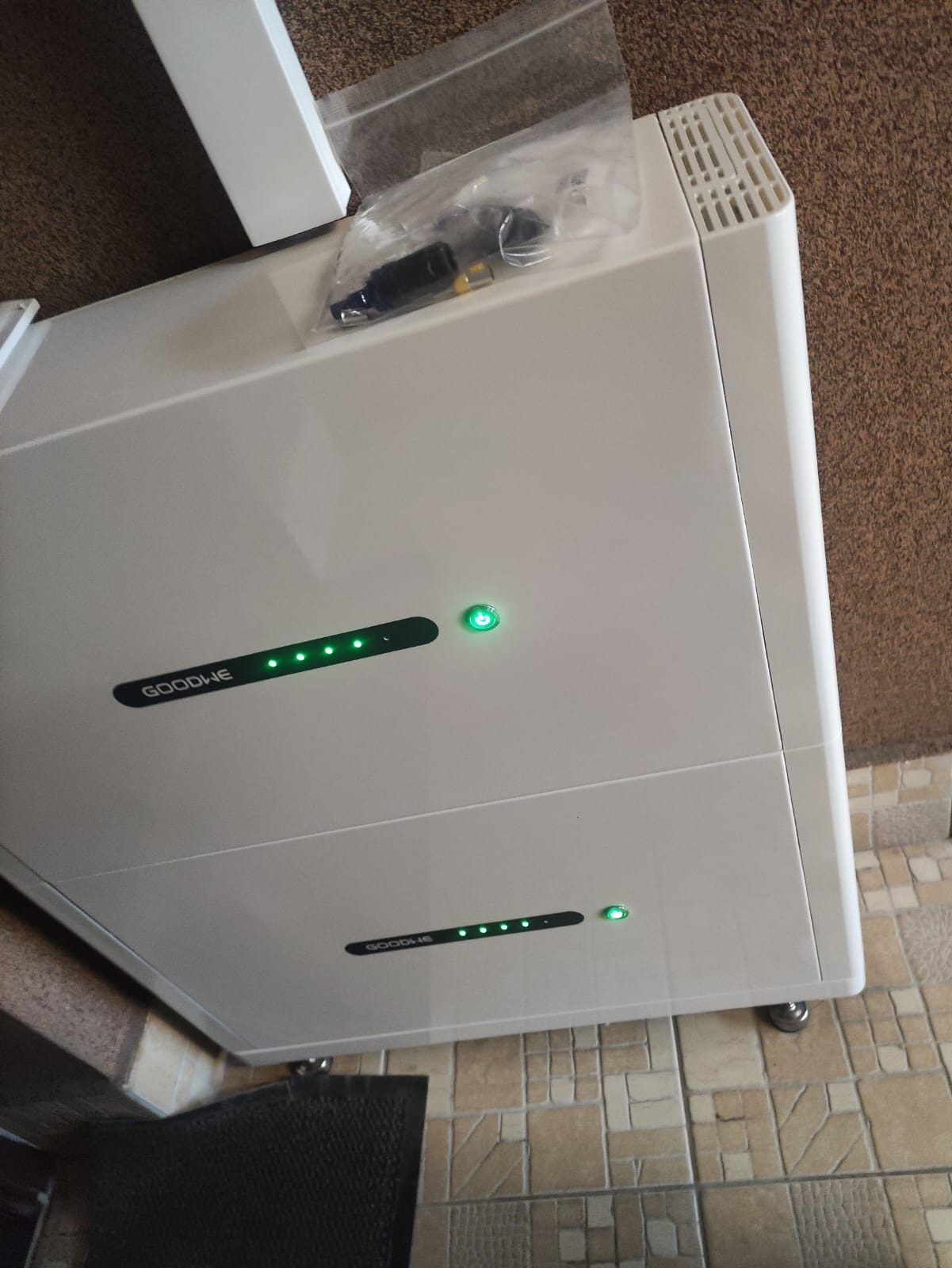 Biały magazyn energii GoodWe z zielonymi diodami LED, widoczne akcesoria w folii. Urządzenie stoi na kafelkach, obok ściana z fakturą. Ujęcie z góry.