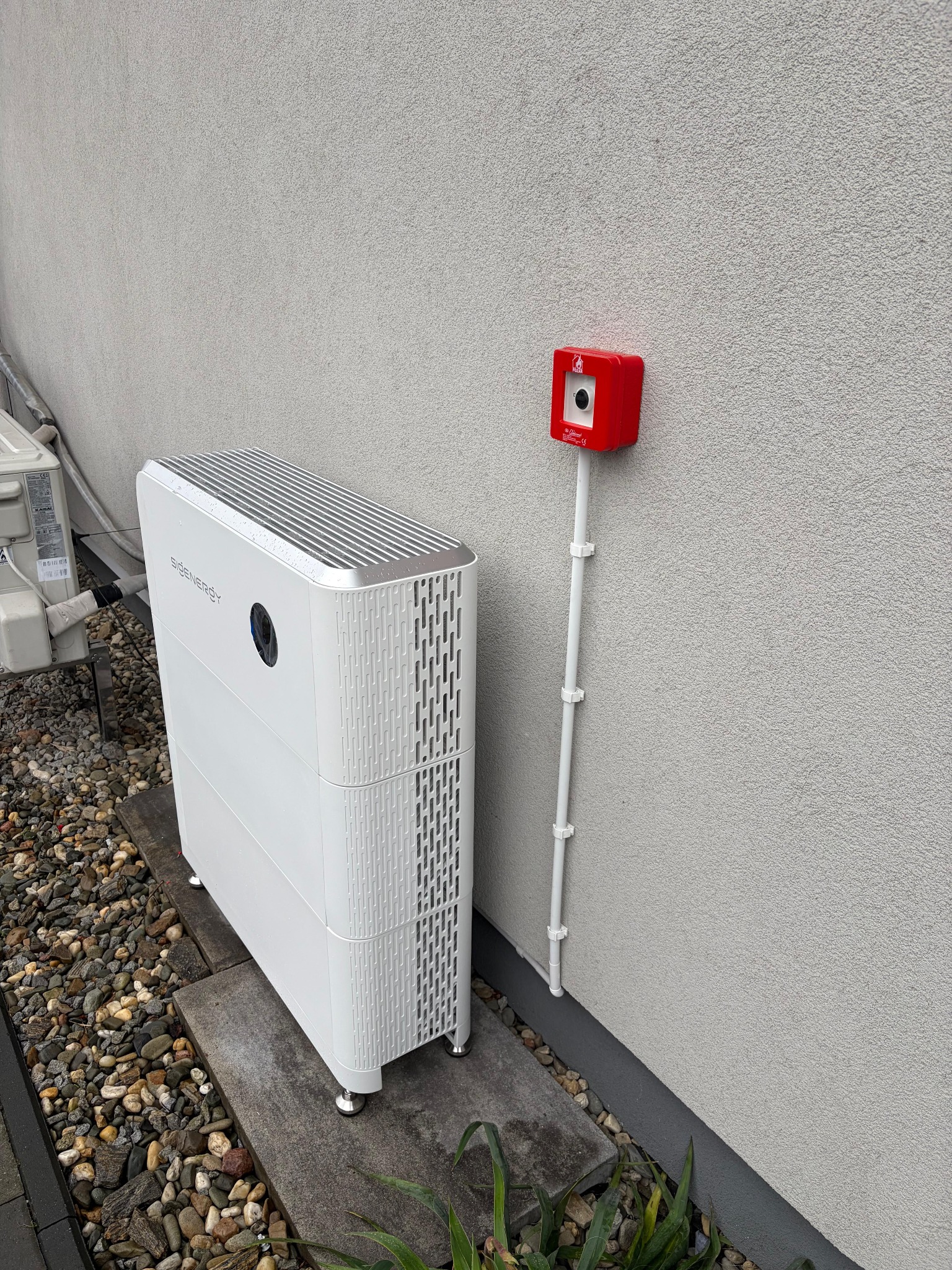 Białe, nowoczesne urządzenie do gromadzenia energii SIDENERGY, stojące na szarych płytach przy jasnej ścianie budynku, z czerwonym rozłącznikiem i kamieniami.