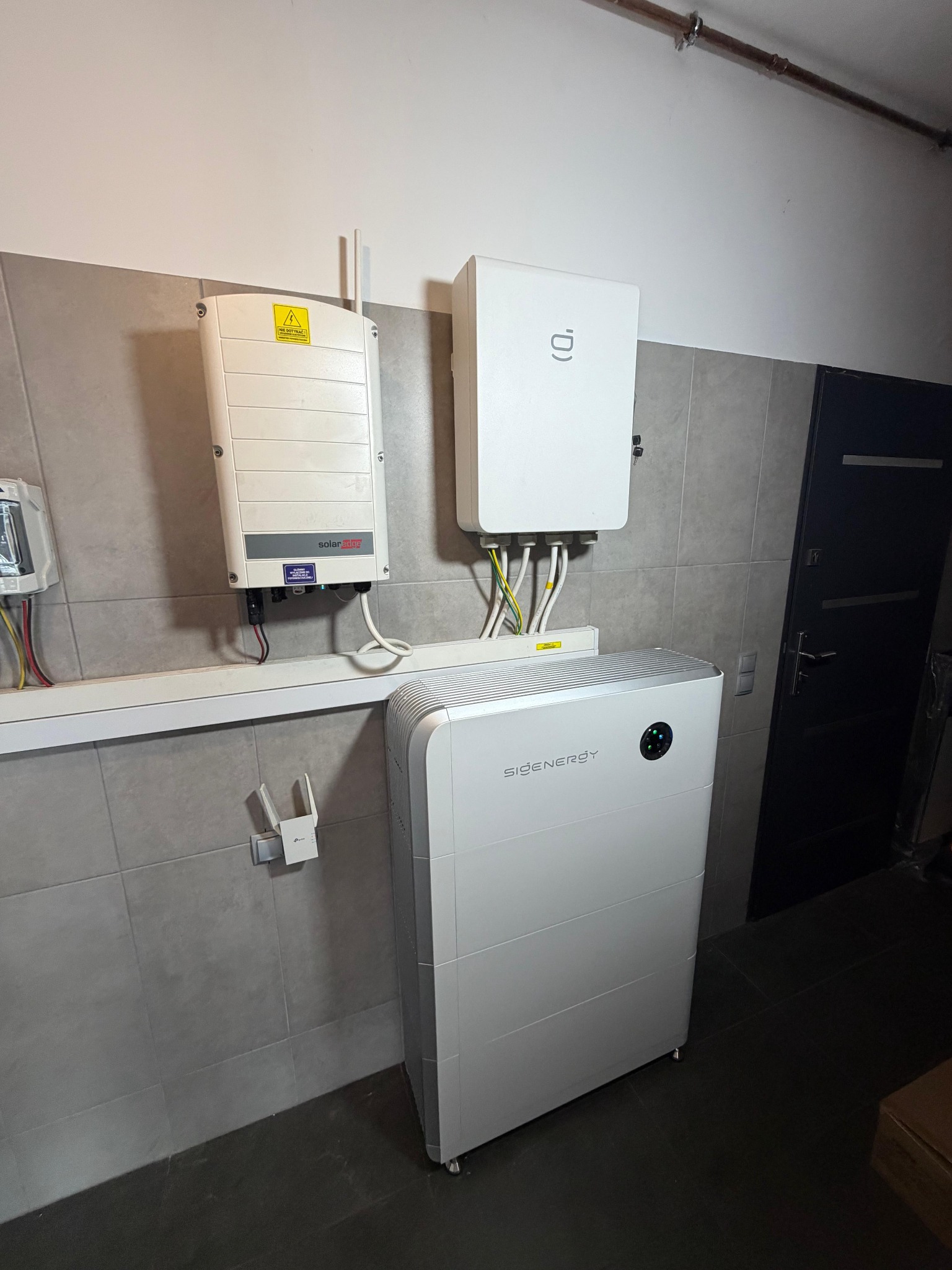 Biały inwerter SolarEdge i pionowy magazyn energii SIDENERGY, profesjonalnie zamontowane na szarej ścianie z dużych płytek, z widocznym okablowaniem i wzmacniaczem Wi-Fi.