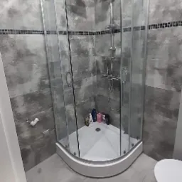 Szklana kabina prysznicowa narożna w szaro-białej łazience z mozaikowym pasem na ścianie, widoczne kosmetyki wewnątrz kabiny i fragment toalety.