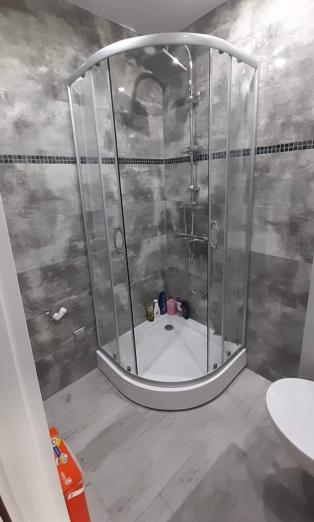 Szklana kabina prysznicowa narożna w szaro-białej łazience z mozaikowym pasem na ścianie, widoczne kosmetyki wewnątrz kabiny i fragment toalety.