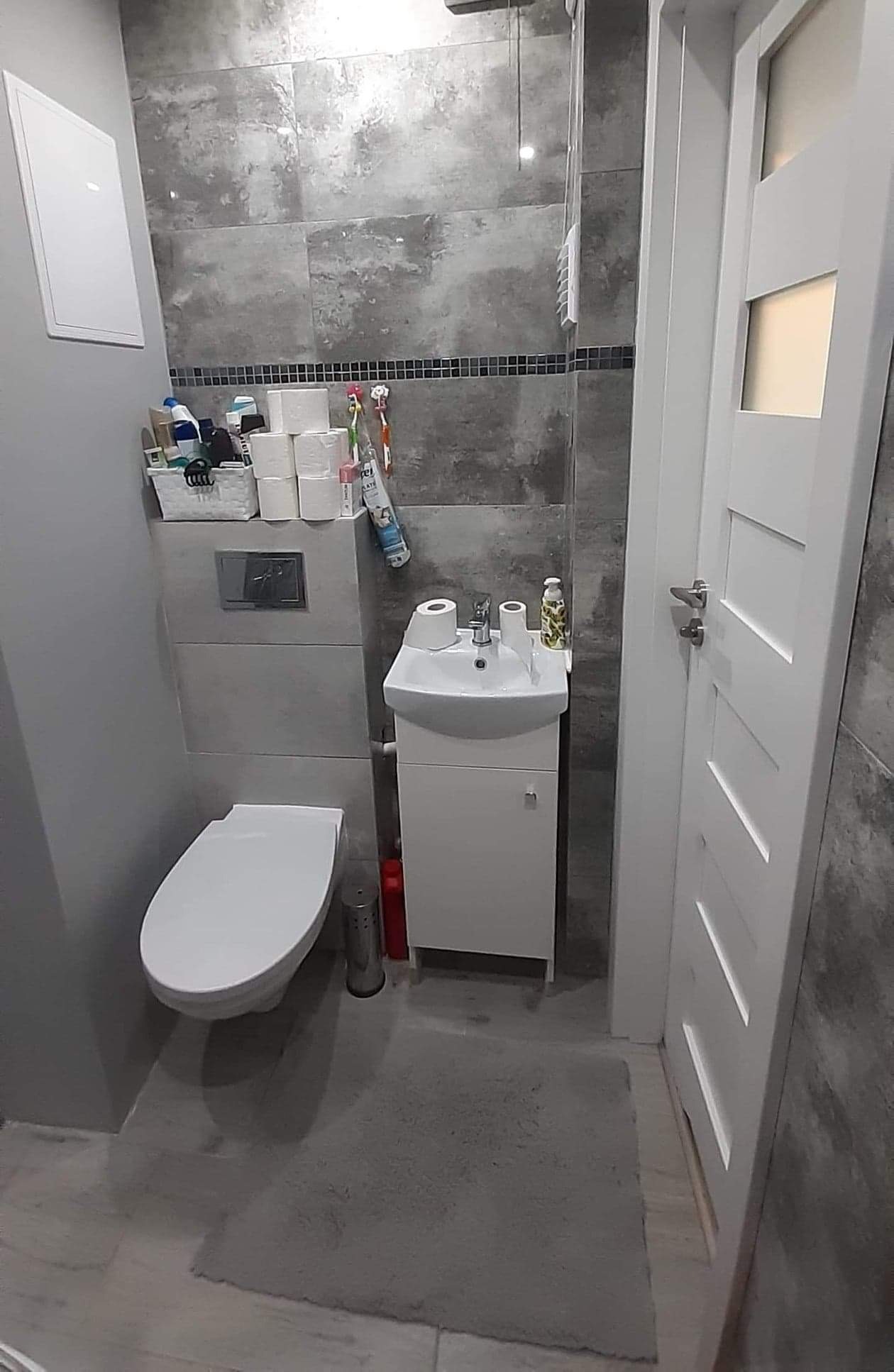 Szara łazienka z umywalką na szafce, toaletą i dużymi płytkami imitującymi beton na ścianach, akcentem są czarne, drobne płytki w pasie pod sufitem, a na umywalce leży rolka papieru toaletowego.