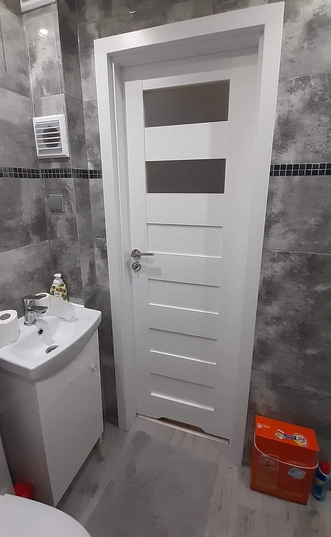 Białe drzwi z przeszkleniami w łazience wykończonej szarymi płytkami, widoczna umywalka z szafką, wentylator i akcesoria do sprzątania.