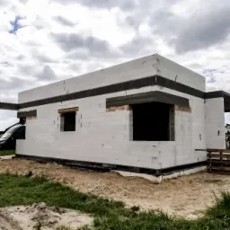 Surowy stan domu murowanego z betonu komórkowego, widoczne otwory okienne i drzwiowe, zbrojenie nadproży, paleta budowlana w tle.