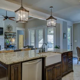 Kuchnia z wyspą, granitowym blatem, białym zlewem farmerskim i ciemnymi drewnianymi szafkami, otwarta na salon z kominkiem i telewizorem