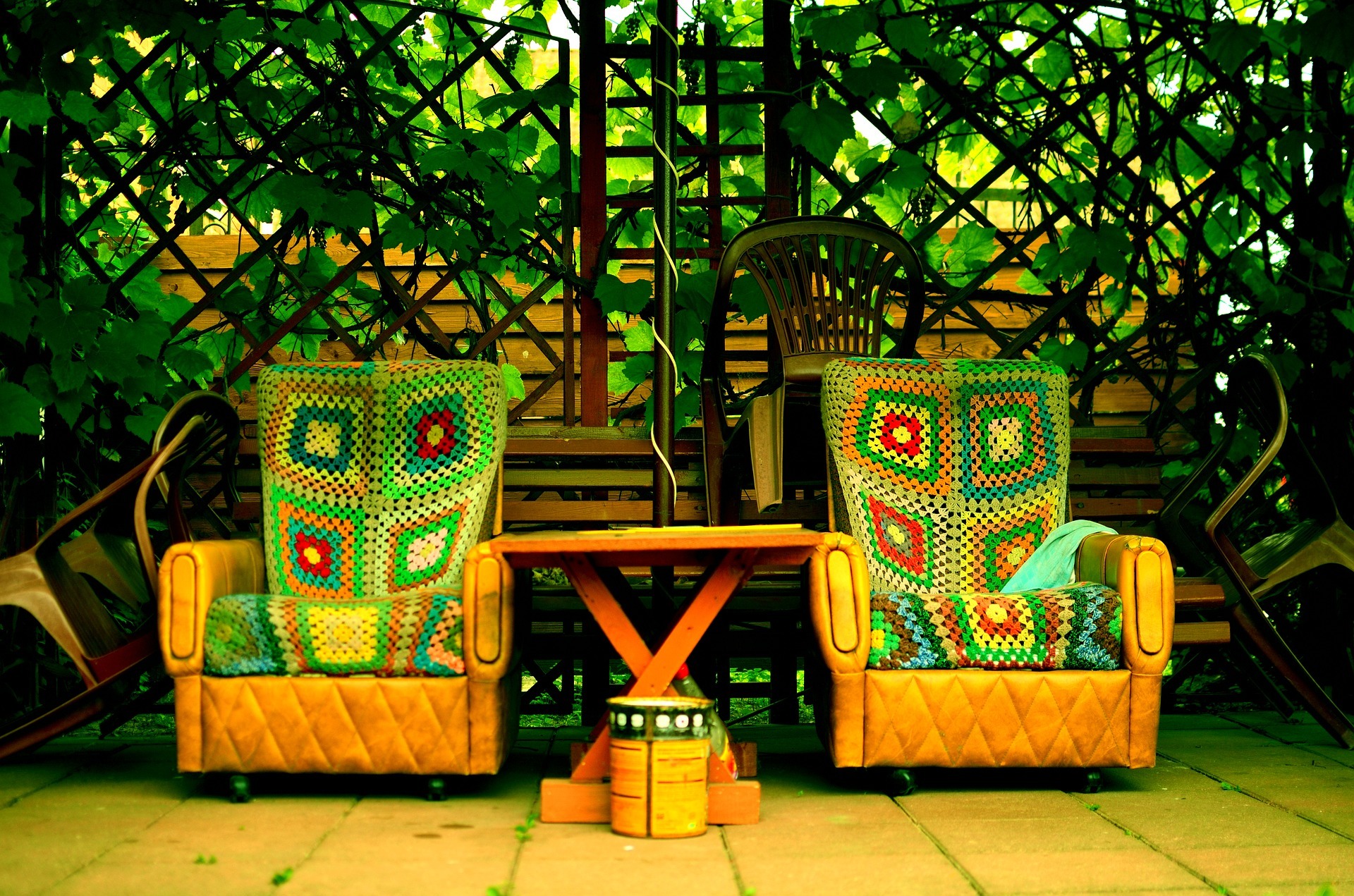 Dwa fotele w stylu retro obite kolorową patchworkową tkaniną, ustawione na tarasie pod drewnianą pergolą porośniętą zielonymi liśćmi winorośli, z małym drewnianym stolikiem pośrodku i pomalowaną...