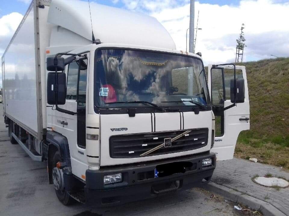 Biała ciężarówka Volvo z otwartymi drzwiami kierowcy zaparkowana na poboczu, widoczne odbicie chmur w szybie.