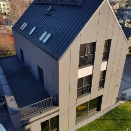 Nowoczesny budynek mieszkalny z grafitową blachą trapezową na dachu i elewacją w odcieniach szarości, widziany z góry w słoneczny dzień.