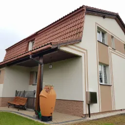 Wykończony dom jednorodzinny z ocieploną elewacją w odcieniach beżu i brązu, z czerwoną dachówką, widok z zewnątrz na fragment elewacji z dekoracyjnym wzorem, ławką i ozdobnym zbiornikiem...