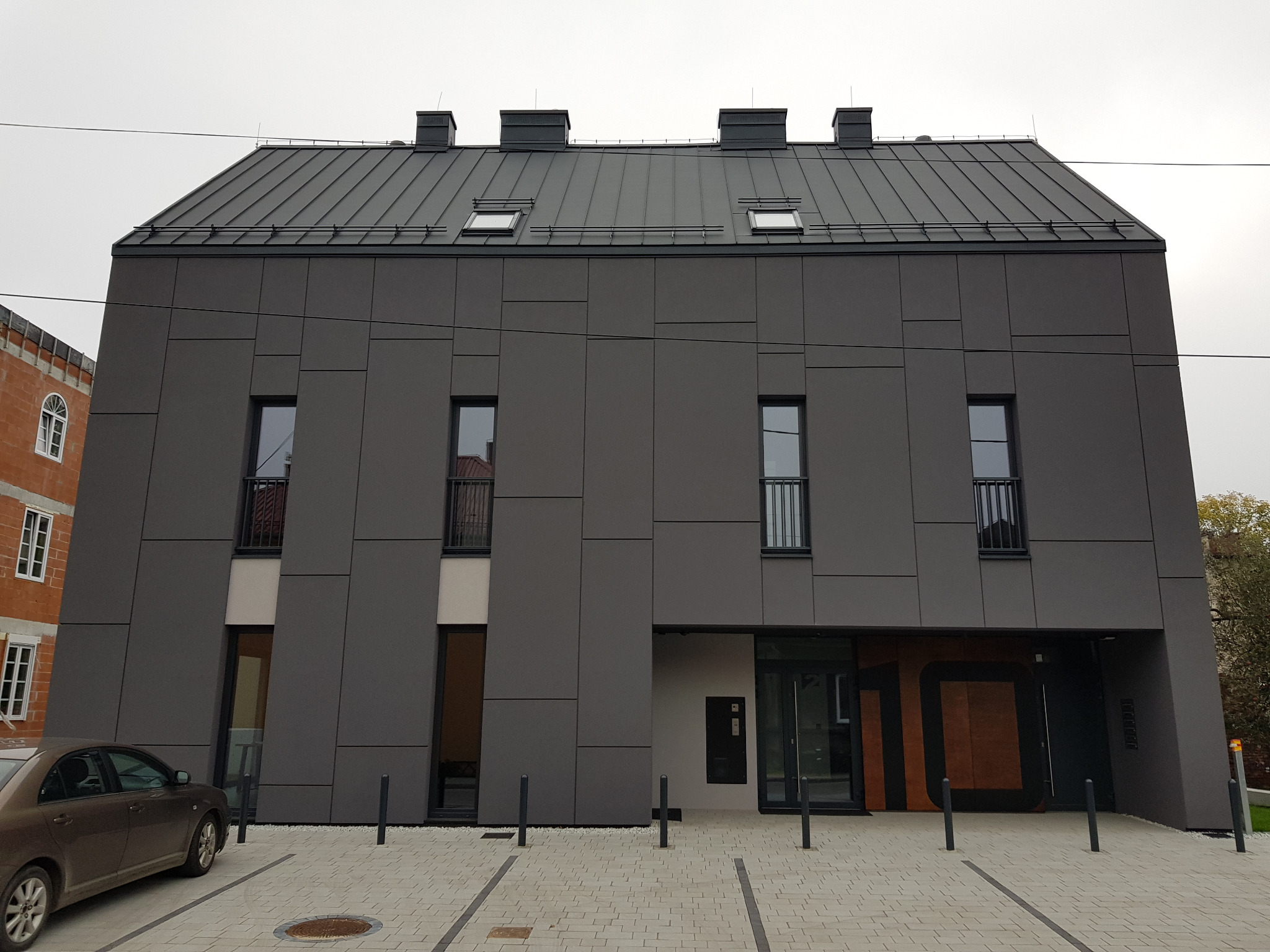Nowoczesny budynek z szarą elewacją panelową i ciemnym dachem z blachy, widok z zewnątrz na wejście z numerem 10, samochód zaparkowany po lewej stronie.