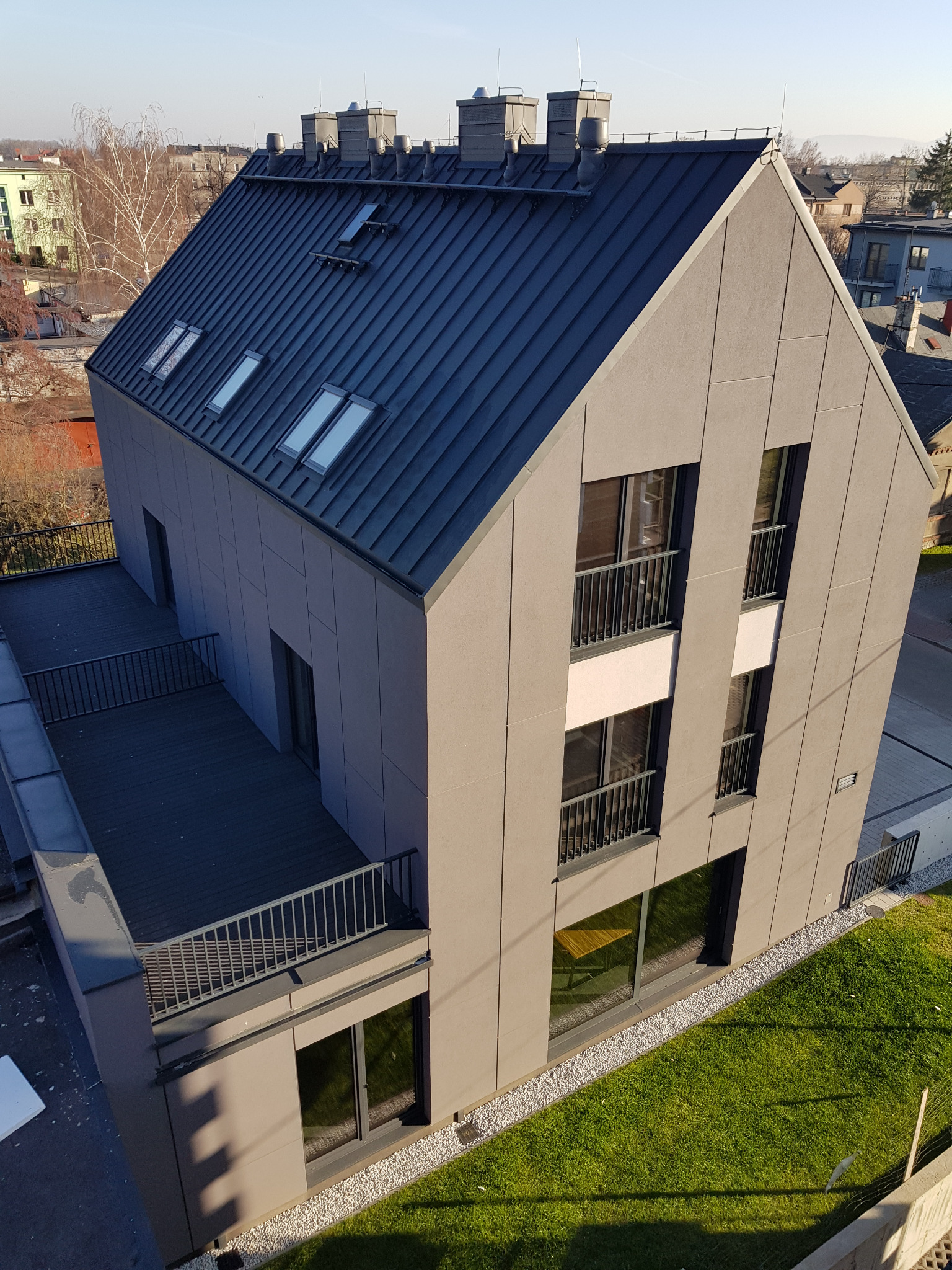 Nowoczesny budynek mieszkalny z grafitową blachą trapezową na dachu i elewacją w odcieniach szarości, widziany z góry w słoneczny dzień.