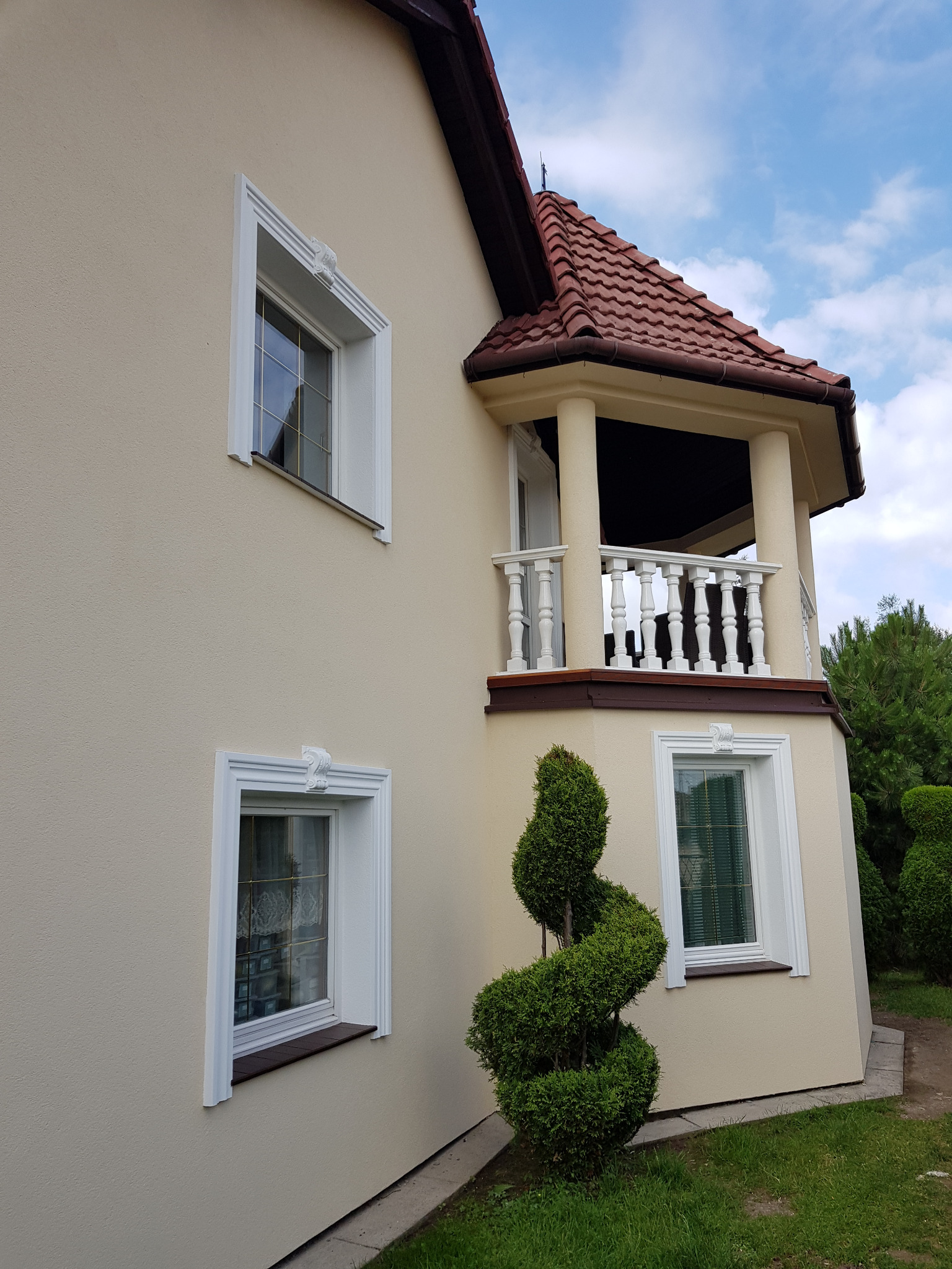 Dom jednorodzinny z beżową elewacją, białymi ramami okiennymi z dekoracyjnymi elementami, balkonem z kolumnami i czerwoną dachówką, oraz spiralnie uformowanym krzewem.