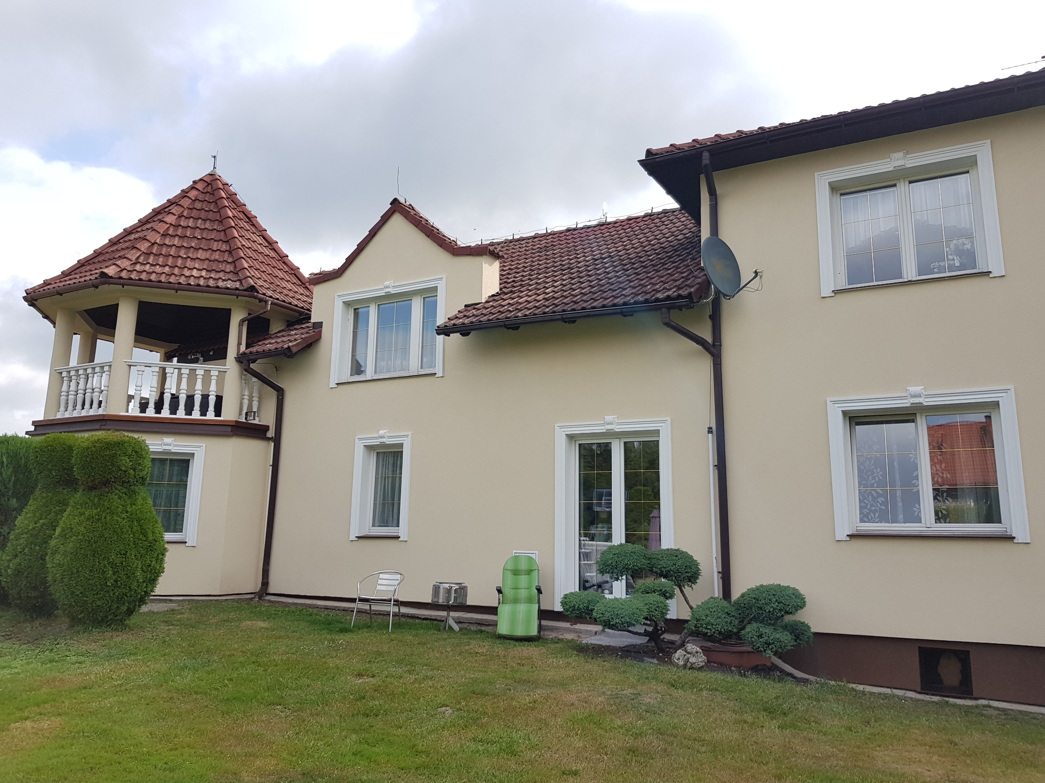 Dwupiętrowy dom z beżową elewacją, brązowym dachem z dachówką i balkonem z balustradą, otoczony zielonym trawnikiem i ozdobnymi krzewami.