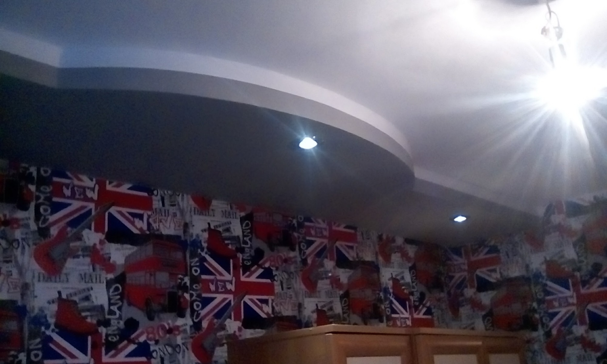 Wykończony fragment pokoju z podwieszanym sufitem z wbudowanym oświetleniem punktowym i ścianą pokrytą tapetą z motywami brytyjskimi, widoczny fragment mebla pod ścianą.