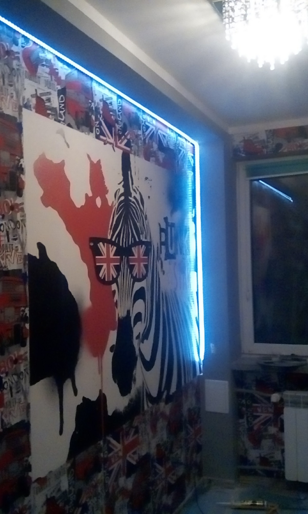 Ściana pokoju z fototapetą przedstawiającą zebrę w okularach z motywem flagi brytyjskiej, podświetlona niebieską listwą LED, na tle tapety z motywem flagi UK.