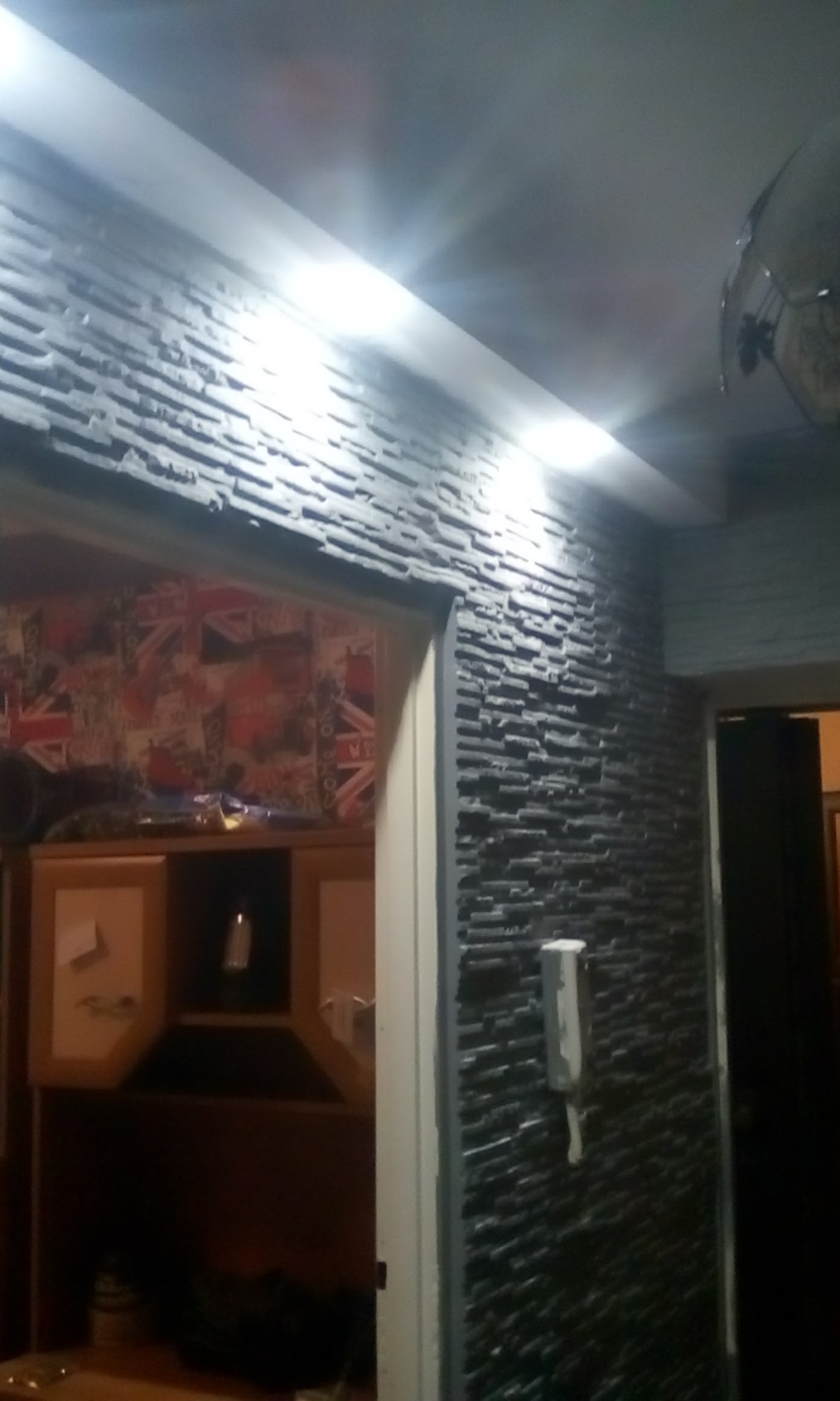 Ściana wykończona szarą okładziną imitującą kamień, oświetlona listwą LED podsufitową, widok na wnętrze z tapetą w motywy brytyjskie i meble w tle.