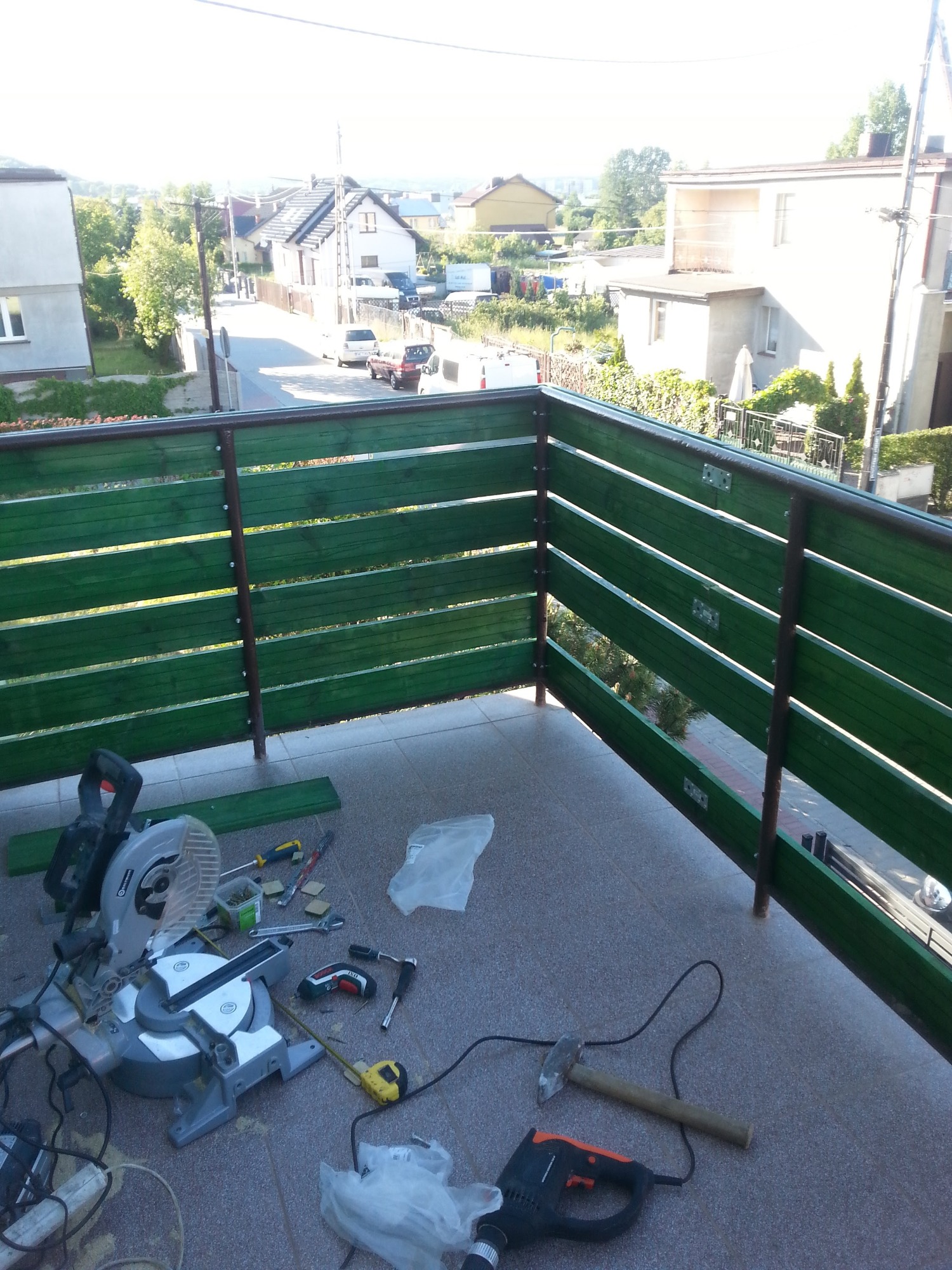 Montaż zielonej, drewnianej balustrady balkonowej z widokiem na zabudowę jednorodzinną, narzędzia pracy rozłożone na płytkach balkonu: ukośnica, wiertarka, młotek, miarka.