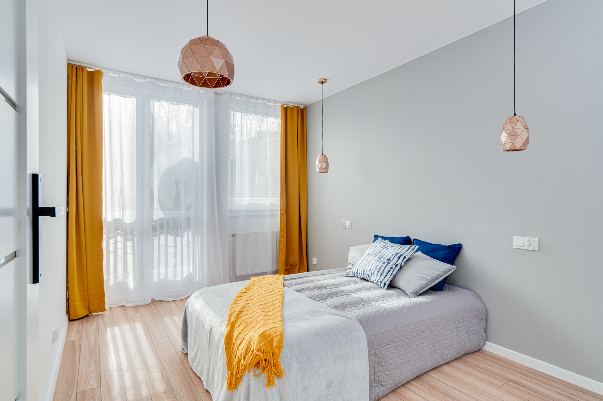 Jasna sypialnia z łóżkiem, żółtym pledem i zasłonami, drewnianą podłogą oraz geometrycznymi lampami wiszącymi.