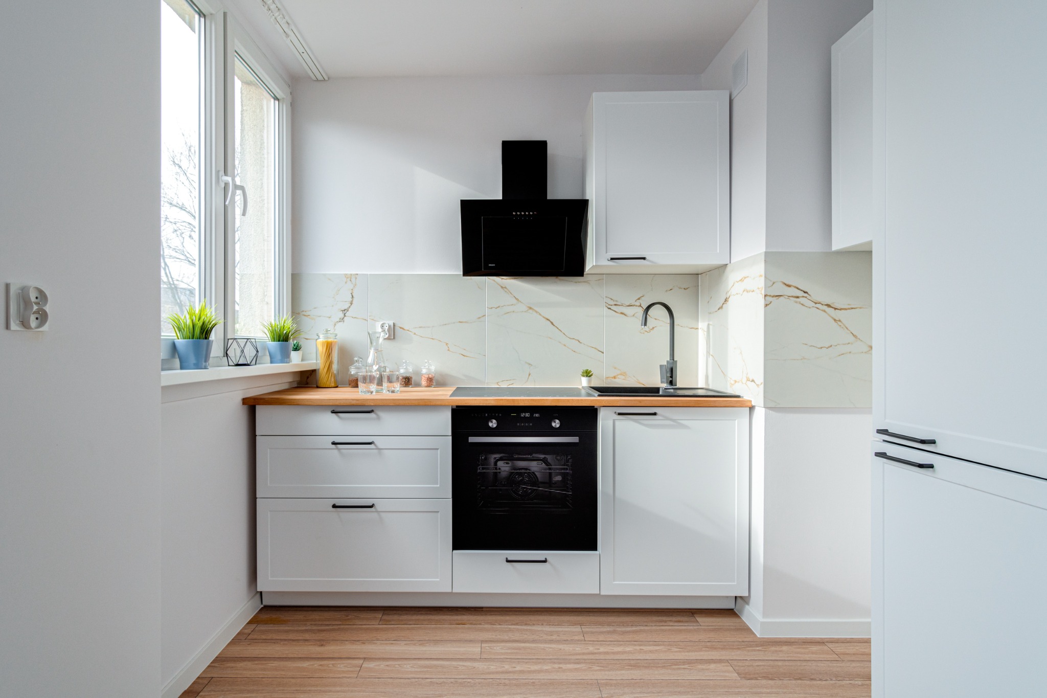Nowoczesna, minimalistyczna kuchnia we Wrocławiu z białymi szafkami, drewnianym blatem i czarnymi akcentami, takimi jak okap i bateria, oświetlona naturalnym światłem wpadającym przez okno...