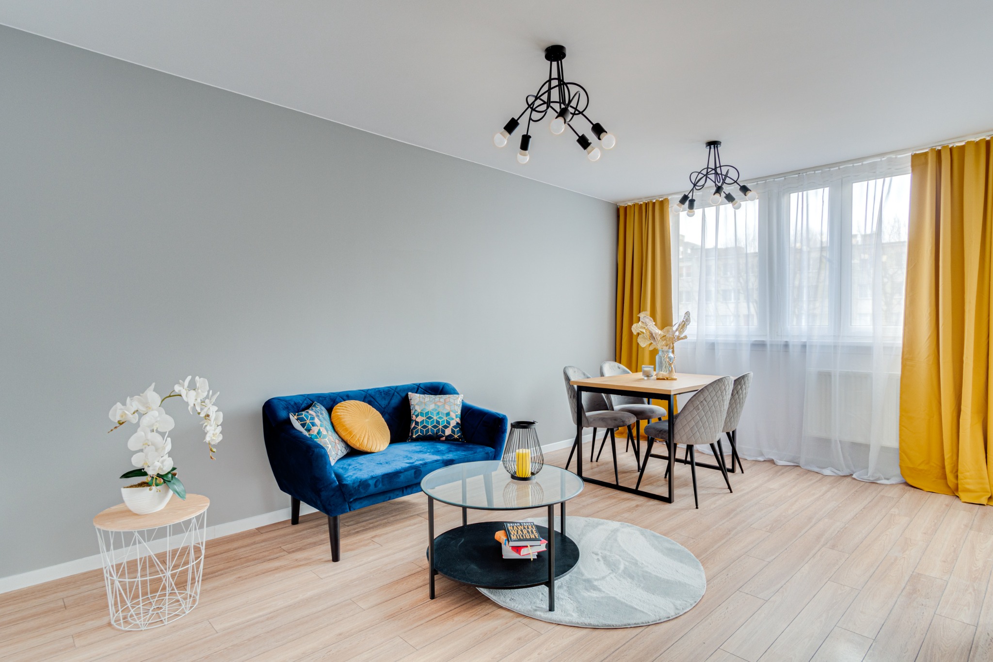 Jasne wnętrze salonu z niebieską sofą, okrągłym stolikiem kawowym z książkami, drewnianym stołem z krzesłami i żółtymi zasłonami przy oknie.