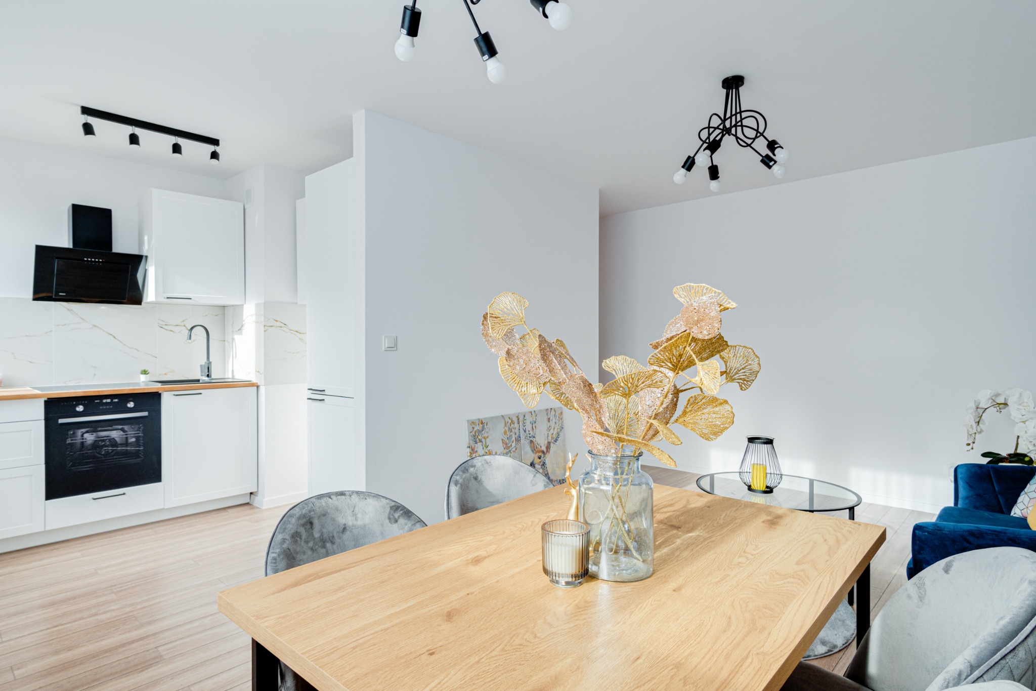 Jasne wnętrze: drewniany stół z dekoracją z liści miłorzębu w szklanym wazonie, w tle biała kuchnia z marmurowym backsplashiem i ciemnymi akcentami, widoczne krzesła obite szarym welurem...
