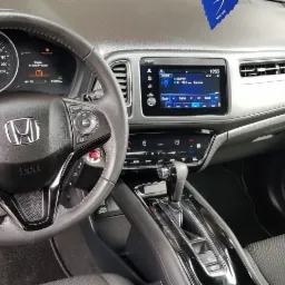 Wnętrze samochodu Honda HR-V z widocznym kokpitem, kierownicą z logo, wyświetlaczem systemu multimedialnego i panelem klimatyzacji; w tle zawieszka zapachowa.