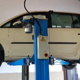 Beżowy samochód typu van na podnośniku w warsztacie, z otwartą maską i widocznymi pracownikami w tle.