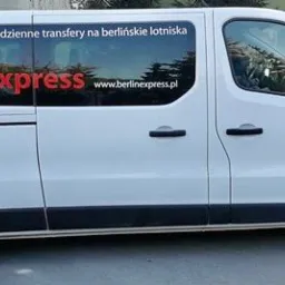 Biały bus z logo BerlinExpress i napisem 'Poznań - Berlin - Poznań, codzienne transfery na berlińskie lotniska' na boku, zaparkowany przed budynkiem.