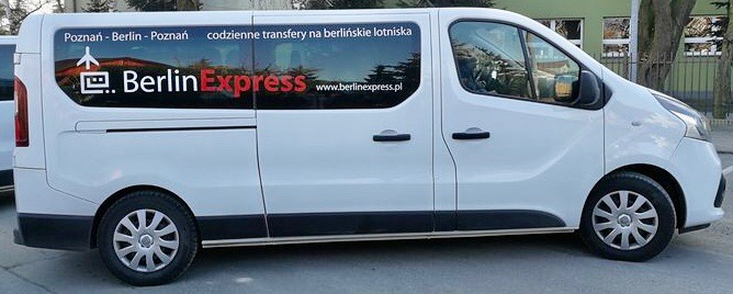 Biały bus z logo BerlinExpress i napisem 'Poznań - Berlin - Poznań, codzienne transfery na berlińskie lotniska' na boku, zaparkowany przed budynkiem.