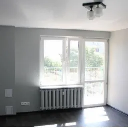 Pomalowane na szaro wnętrze pokoju z grzejnikiem pod oknem balkonowym i lampą sufitową.