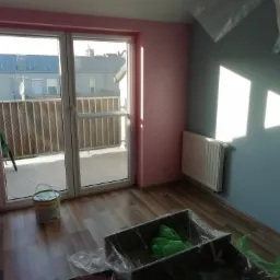 Pokój po remoncie z pomalowanymi ścianami na różowo i niebiesko, widoczny grzejnik, wiadro z farbą, folia malarska i balkon.