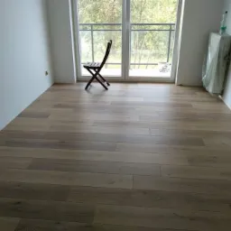 Wykończone wnętrze z nową, jasną podłogą panelową ułożoną wzdłuż pokoju, z widocznym krzesłem i balkonem w tle. Ściany pomalowane na biało, z widocznymi śladami po uszczelnianiu przy podłodze.