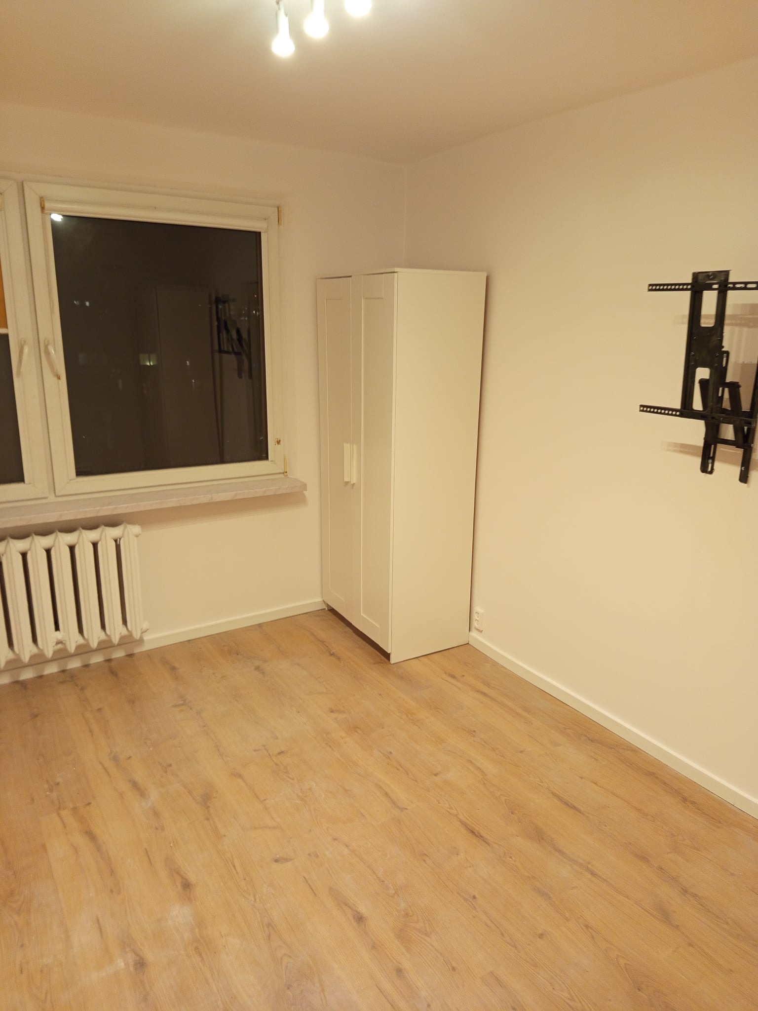 Pusty pokój po remoncie: świeżo pomalowane ściany w odcieniach bieli, jasna podłoga, okno z grzejnikiem i uchwyt na telewizor na ścianie. Minimalistyczne wnętrze.