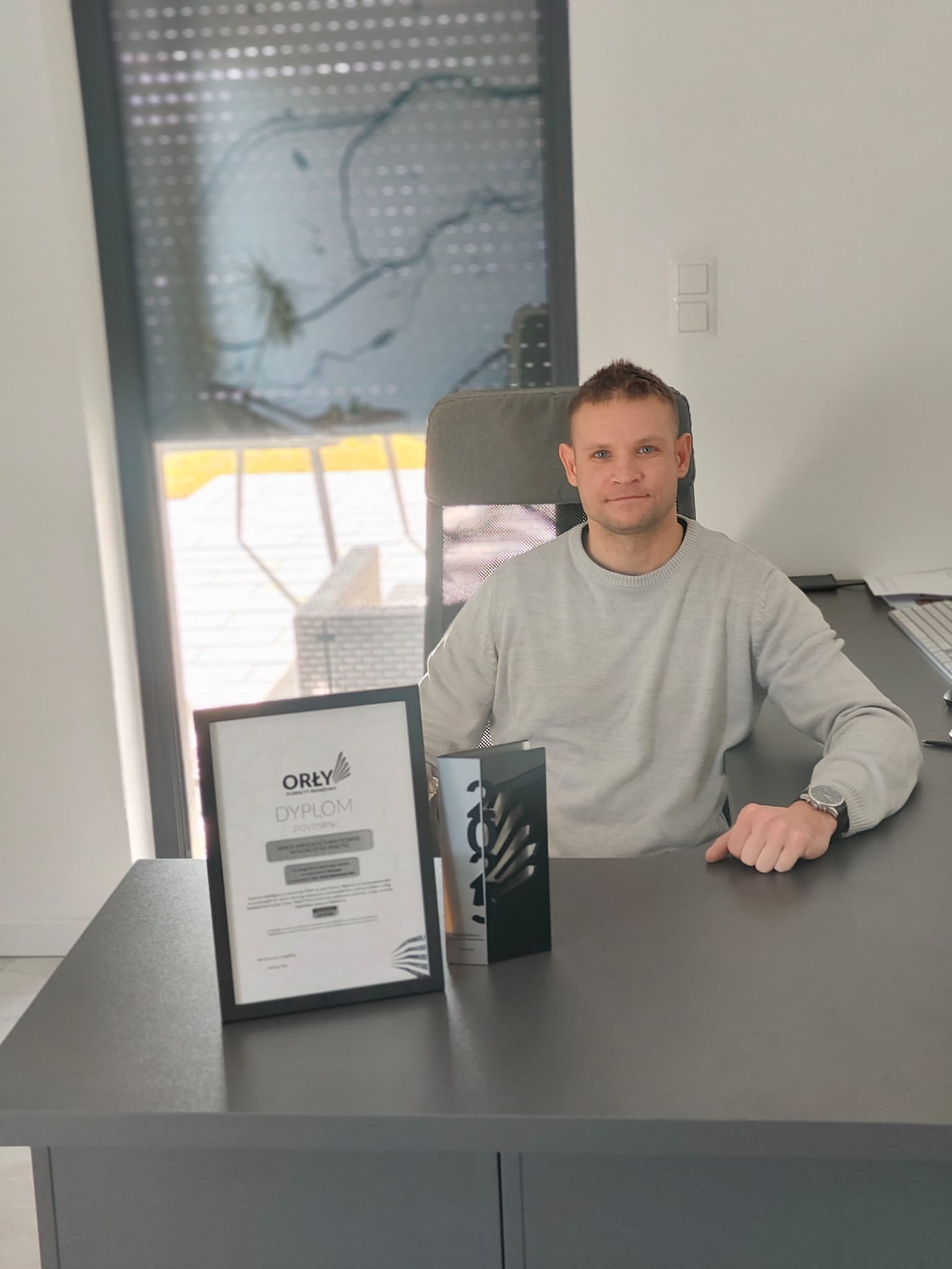 Mężczyzna siedzi za biurkiem z dyplomem 'Orły' w ramce i statuetką. W tle okno z roletami i widok na zewnątrz. Jasne wnętrze, neutralna kolorystyka.